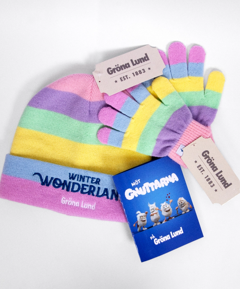 Ett par Gröna Lund Winter Wonderland-vantar och en matchande randig mössa i pastellfärger (rosa, lila, blått, gult och grönt), utlagda tillsammans med en liten blå broschyr för Gnutta-figurerna